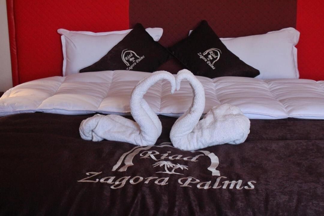 A bed with white bedding, two black pillows with text, and two white towels arranged in a heart shape.