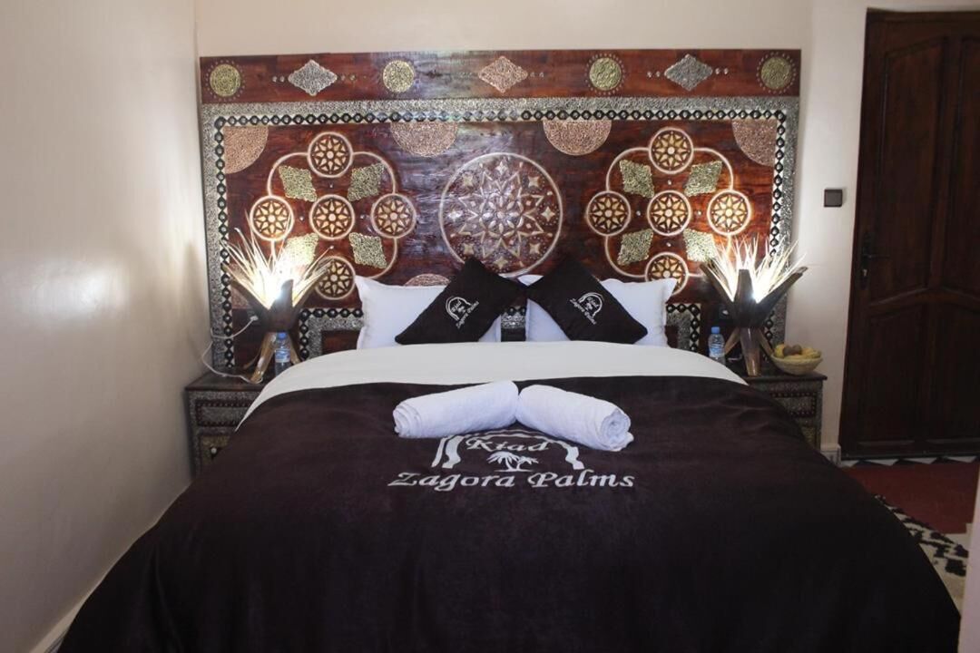 Un lit bien fait, avec une couverture sombre et deux oreillers, une tête de lit décorative ornée de motifs complexes, et une serviette posée sur le lit.