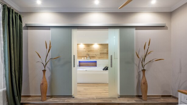 A modern bedroom with a large bed, a bathroom with a wooden vanity, and two vases with dried plants. - Cityhotel Bernburg (Bernburg)