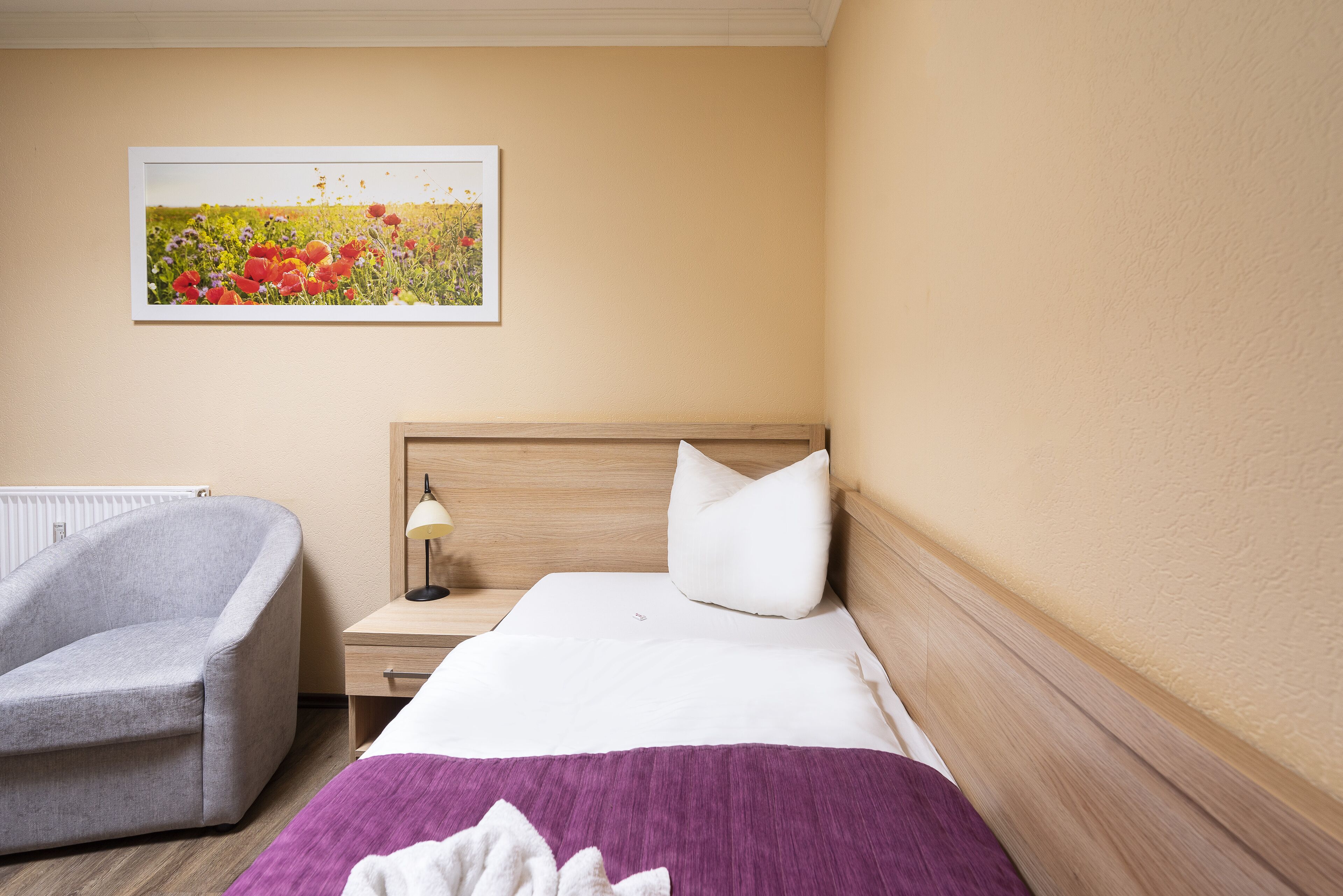 A hotel room with a bed, a chair, a nightstand with a lamp, and a framed picture on the wall.