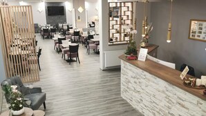 A modern reception area with a wooden reception desk, a seating area with grey sofas and tables, and a dining section with multiple tables and chairs. - Cityhotel Bernburg (Bernburg)