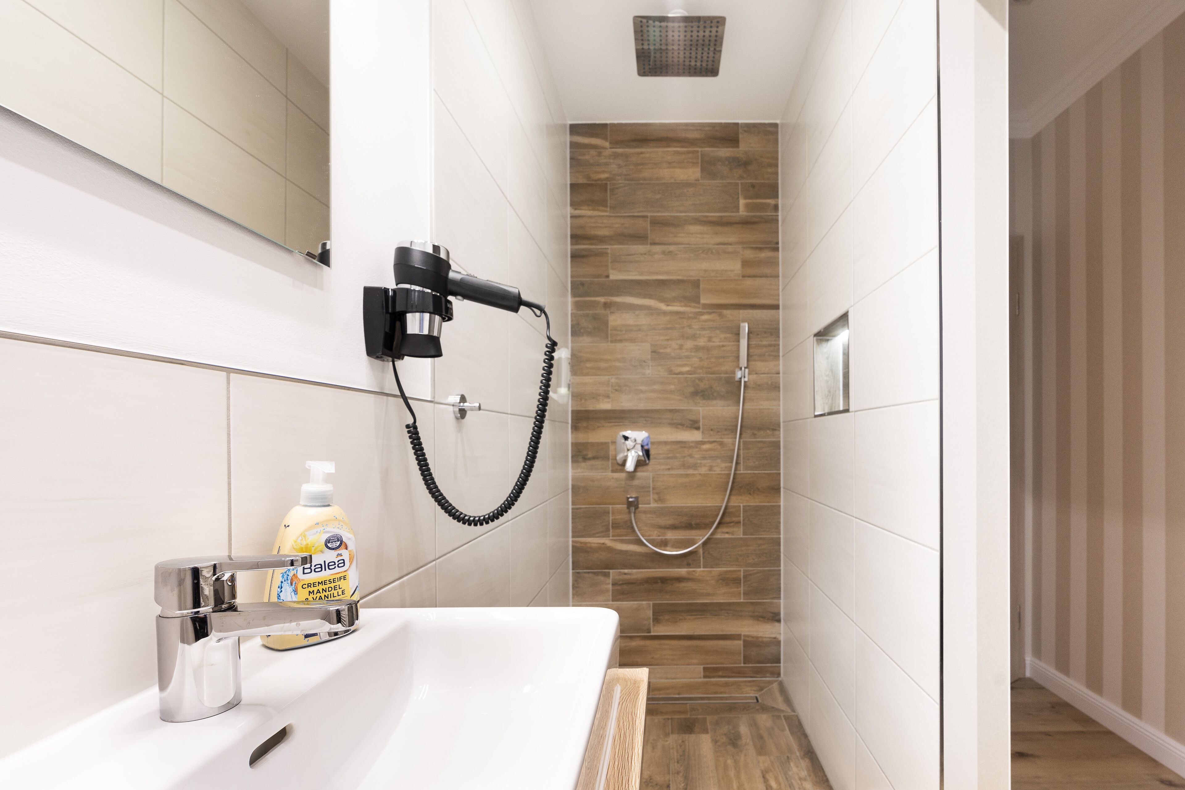 A modern bathroom with a white sink, a handheld showerhead, and a bottle of hand soap.