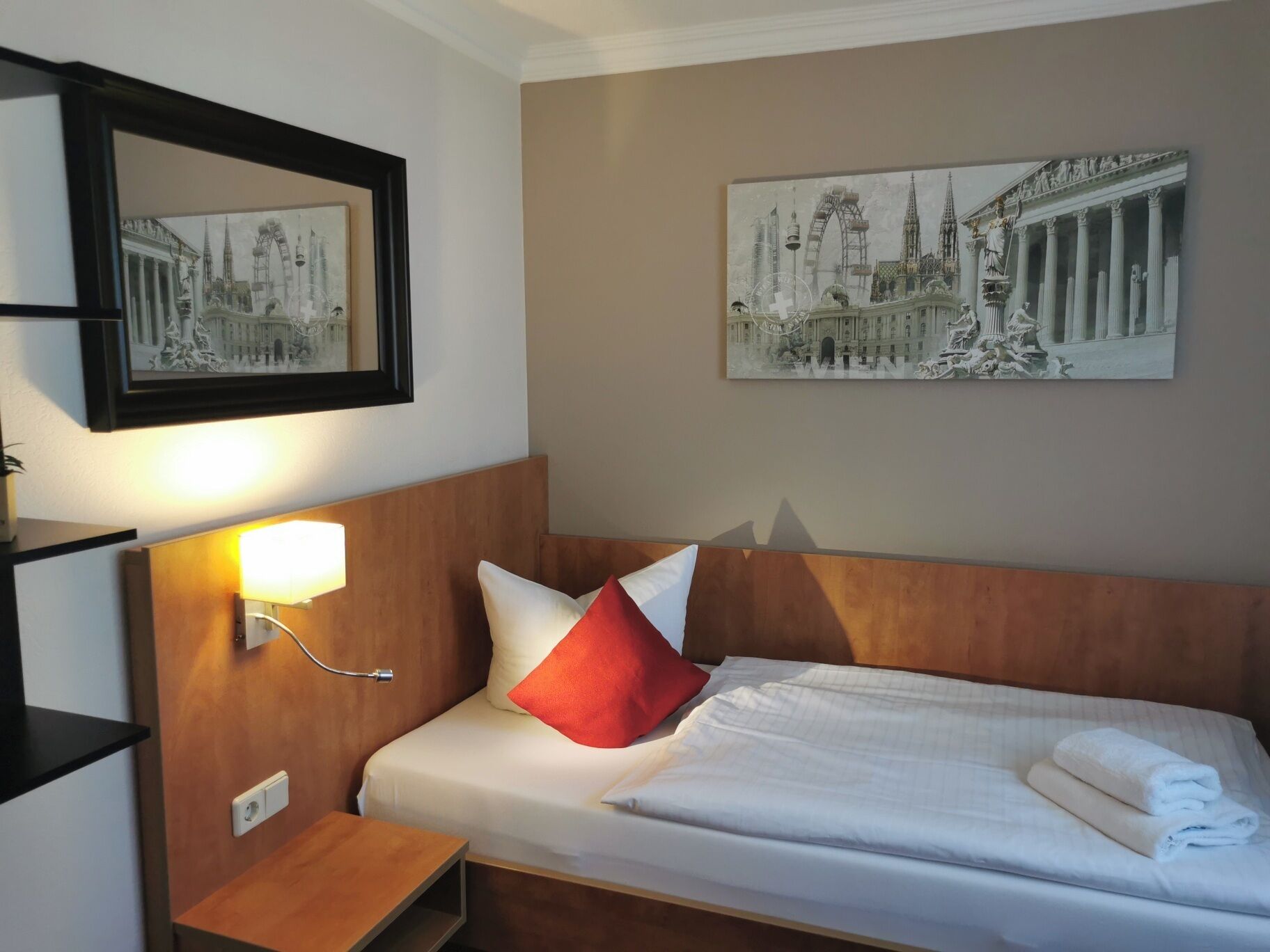 A hotel room with a neatly made bed, a bedside lamp, a small wooden nightstand, and two framed pictures on the wall.