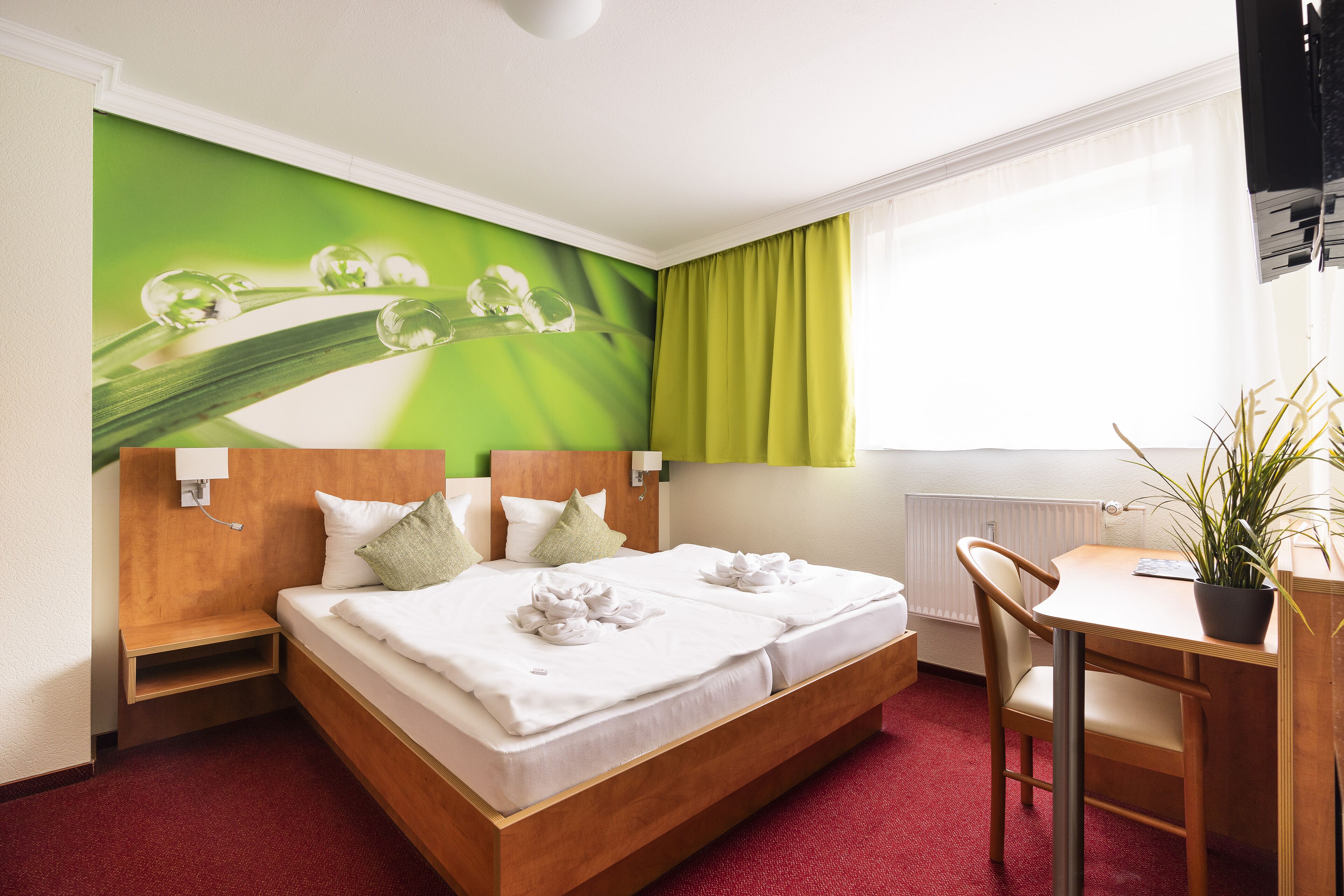 Habitación de hotel con cama, escritorio, silla, planta en maceta y ventana con cortinas.