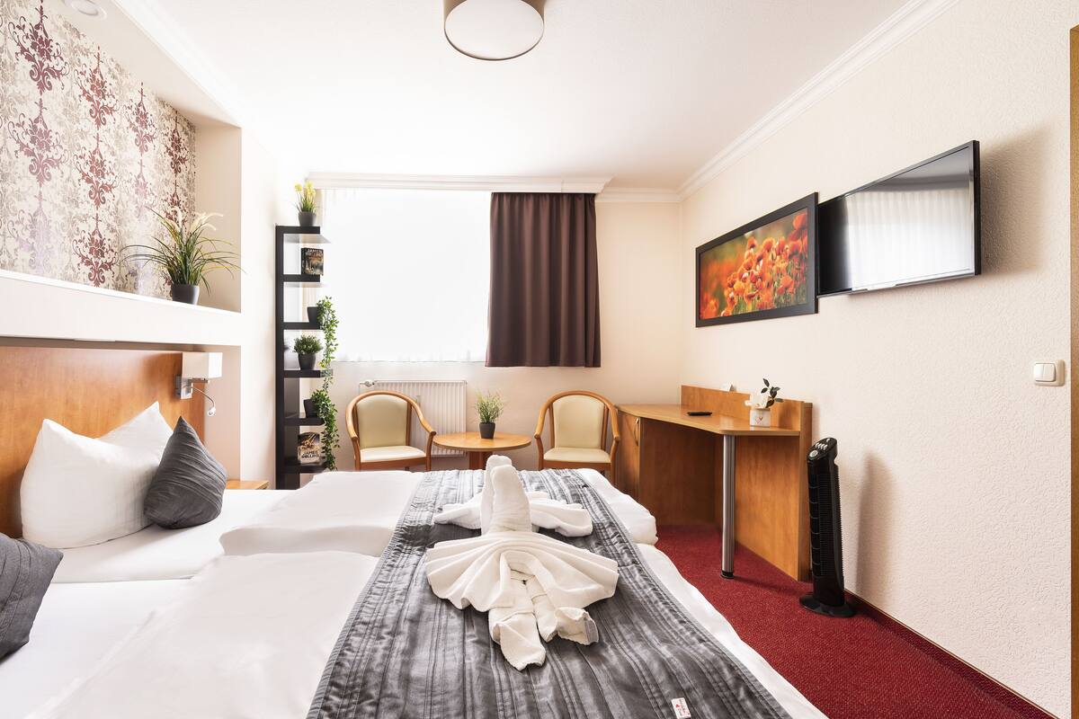 A hotel room with a neatly made bed, a desk, a chair, a small table, and a window with curtains.