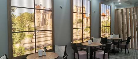 Un comedor moderno con ventanas amplias que ofrecen vistas a un edificio histórico y vegetación. Mesas y sillas dispuestas para comer.