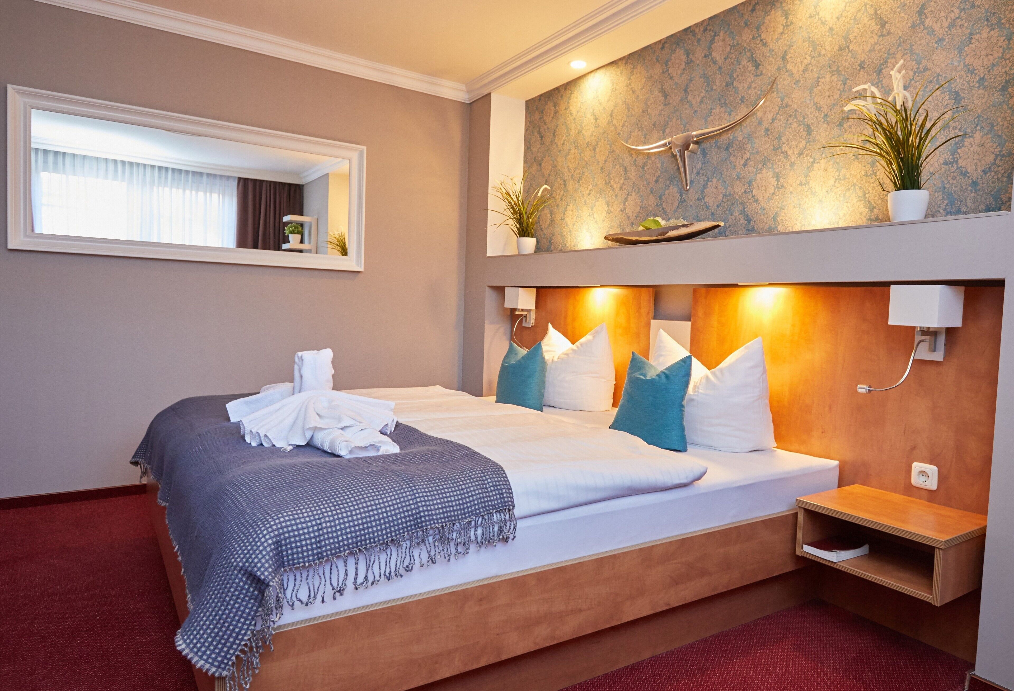 Hotelkamer met een groot bed, een houten hoofdbord, een nachtkastje, een spiegel, een raam met gordijnen en een wandplank met planten.