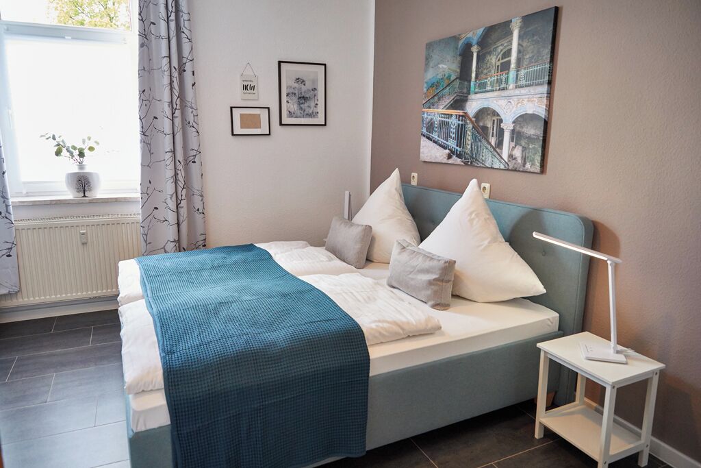 Una cama bien hecha con un edredón verde azulado, una mesita de noche con una lámpara y una ventana con cortinas.