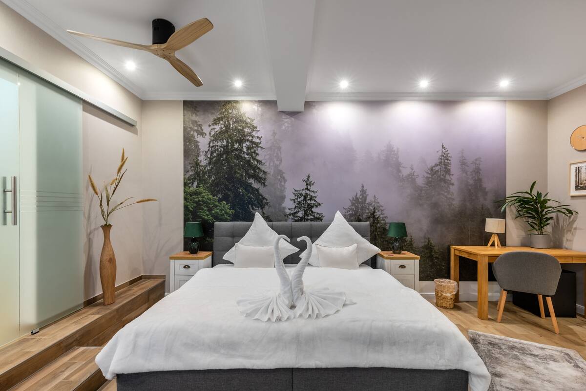 A modern bedroom with a large bed, a ceiling fan, a mural of trees, a desk, and a chair.