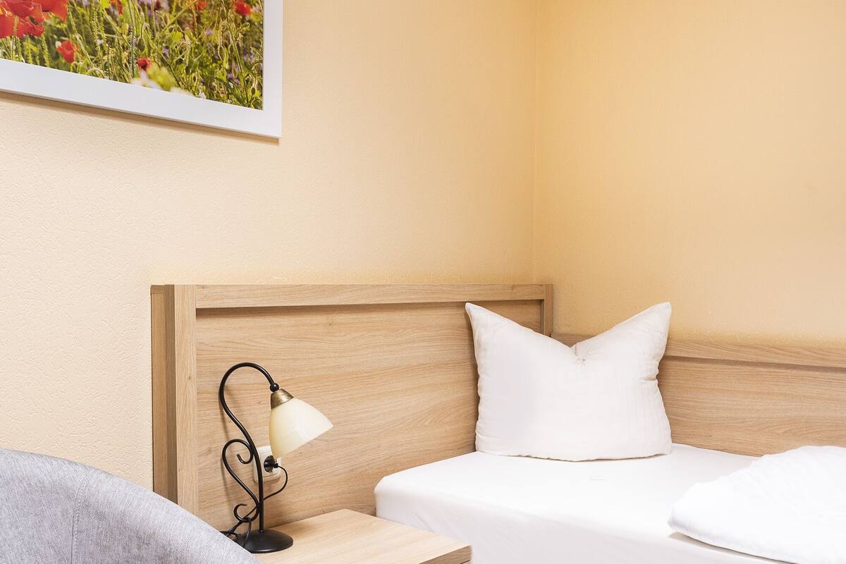 A hotel room with a bed, a nightstand with a lamp, a chair, and a painting on the wall.