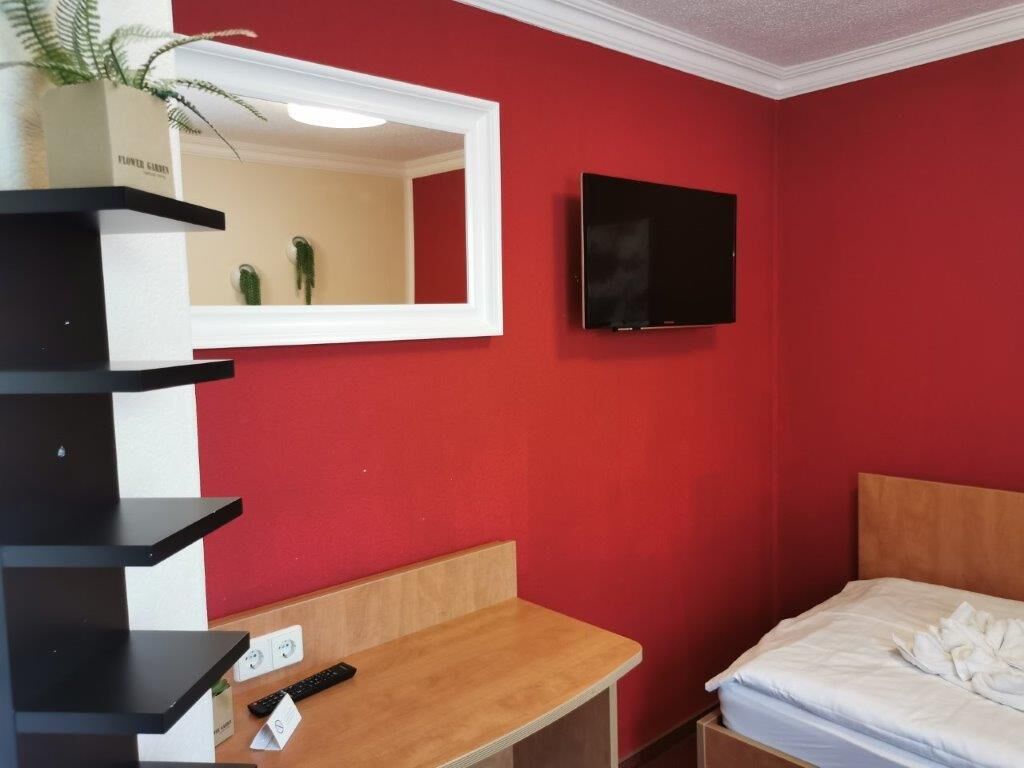 Une pièce avec un mur rouge, un lit avec une literie blanche, un bureau en bois, un miroir, un téléviseur à écran plat et une plante posée sur le bureau.