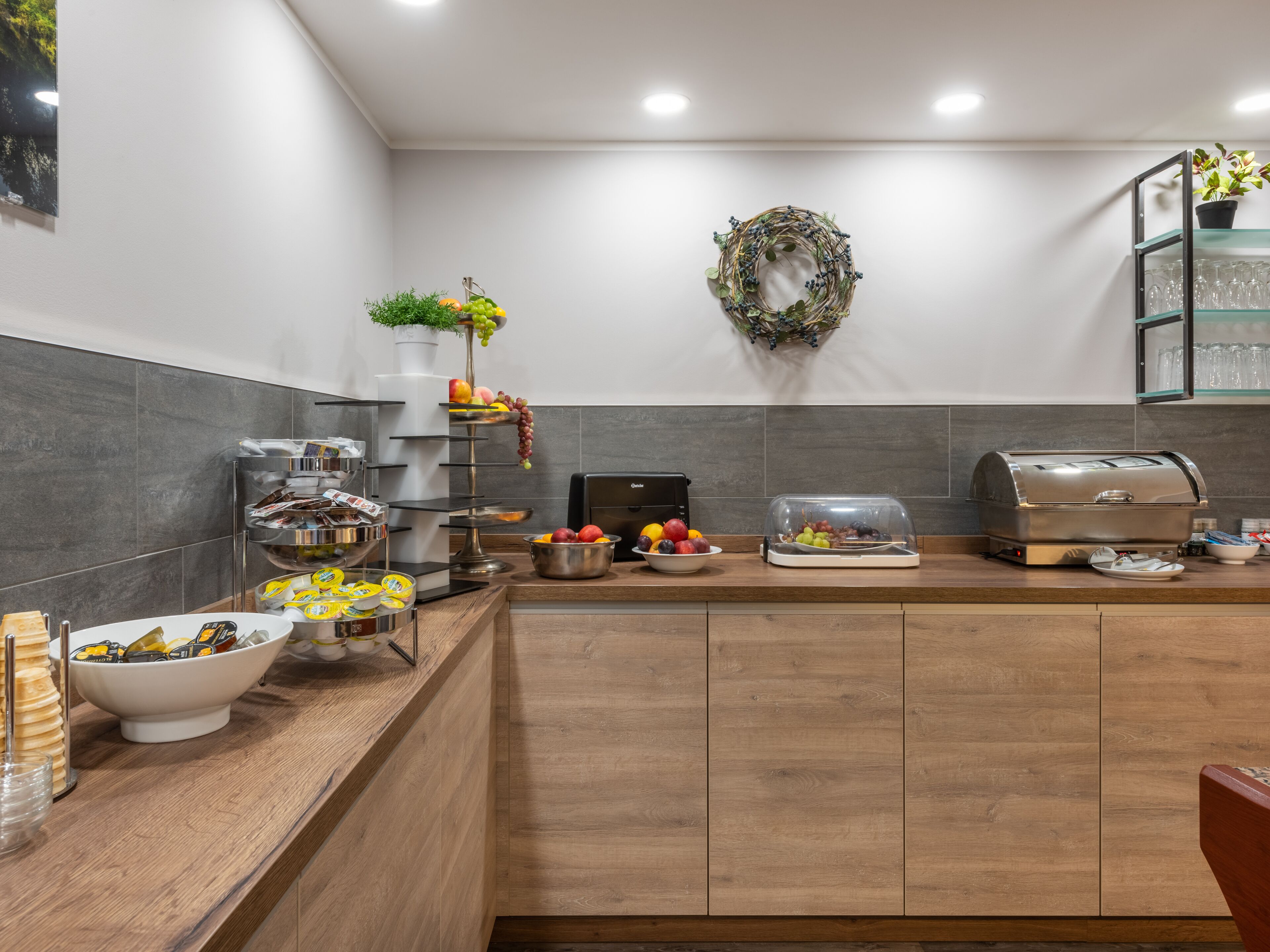 A modern kitchen with a wooden countertop, a coffee station, a fruit display, and a decorative wreath on the wall.