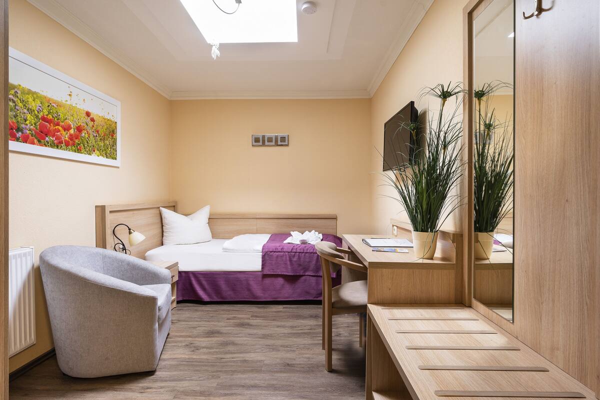 A hotel room with a bed, a desk, a chair, a mirror, a potted plant, and a painting on the wall.