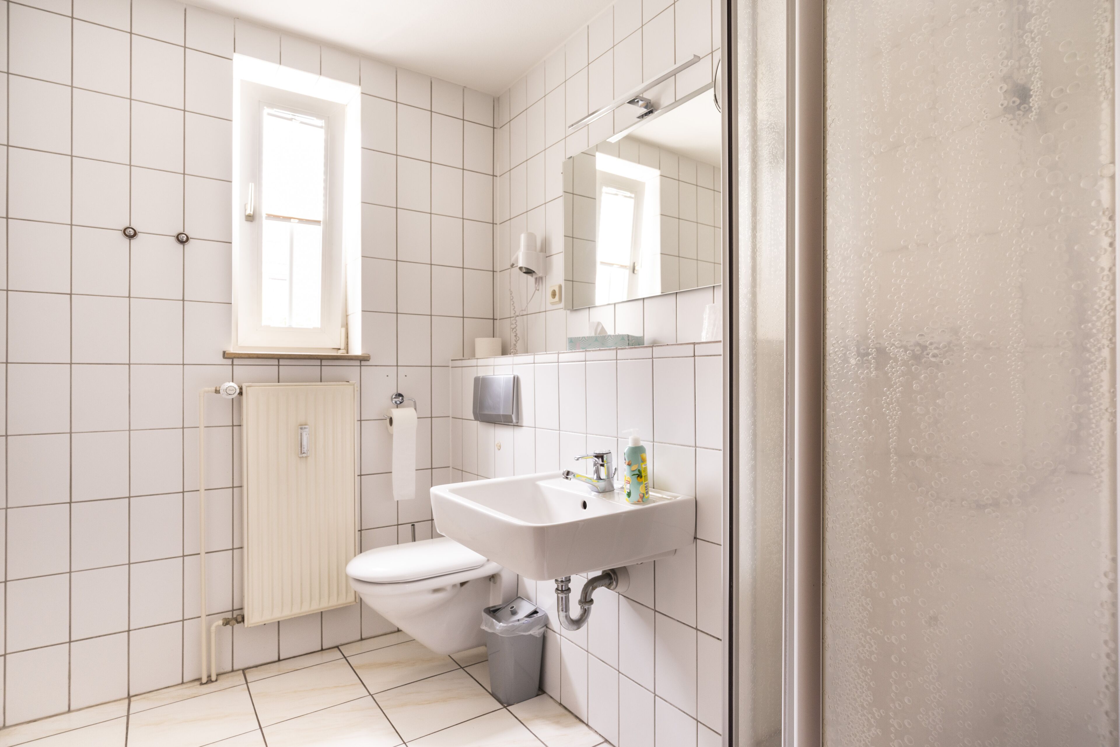 A small, clean bathroom with white tiles, a toilet, a sink, a mirror, and a window.