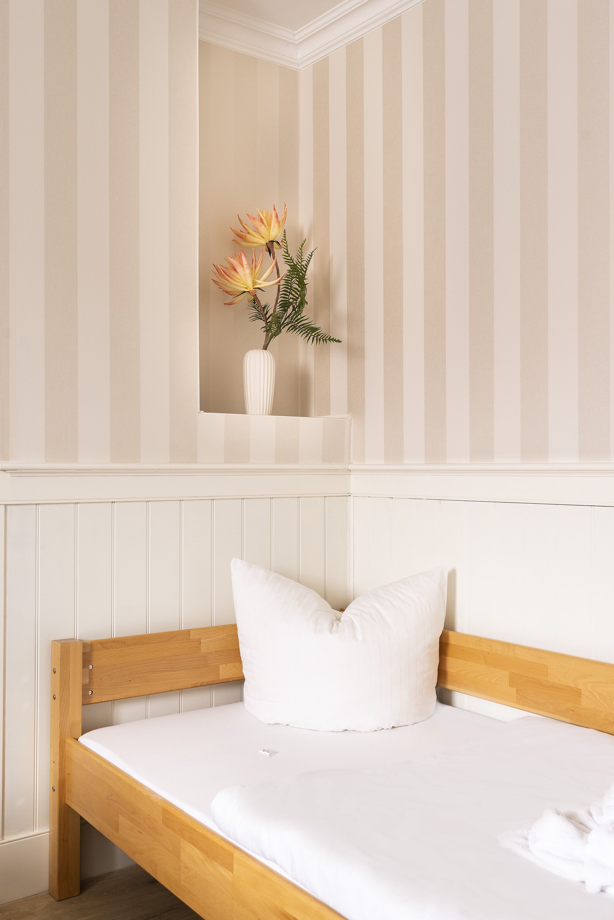 Una cama individual con estructura de madera y ropa de cama blanca, un almohadón blanco y un pequeño florero con flores en la pared de arriba.