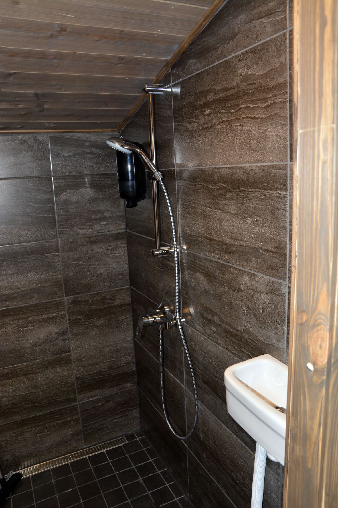 Ducha con cabezal de ducha de mano y grifo empotrado en la pared, inodoro blanco y paredes de madera oscura.