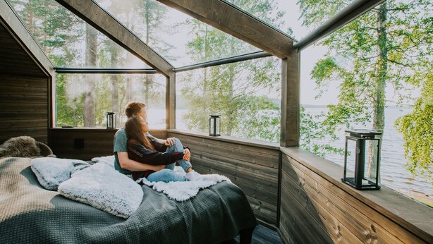 A cozy cabin with a sloped roof, a bed with a fluffy blanket, and a view of a lake through large windows. - Revontuli Resort Glass Igloos (Hankasalmi)