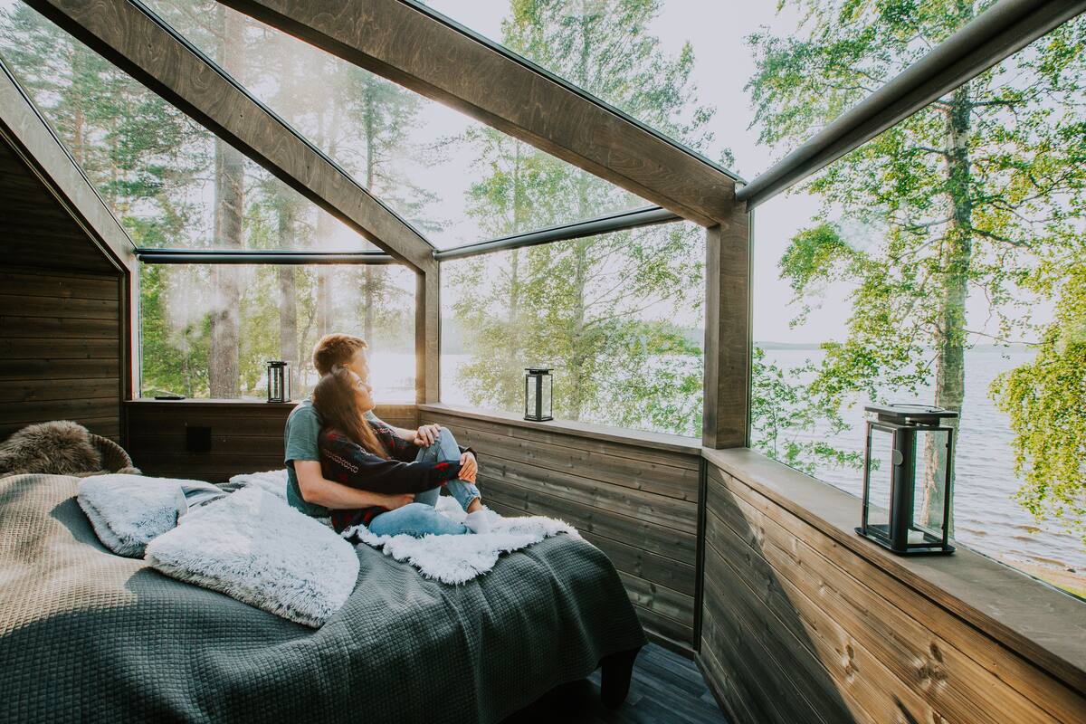 A cozy cabin with a sloped roof, a bed with a fluffy blanket, and a view of a lake through large windows.