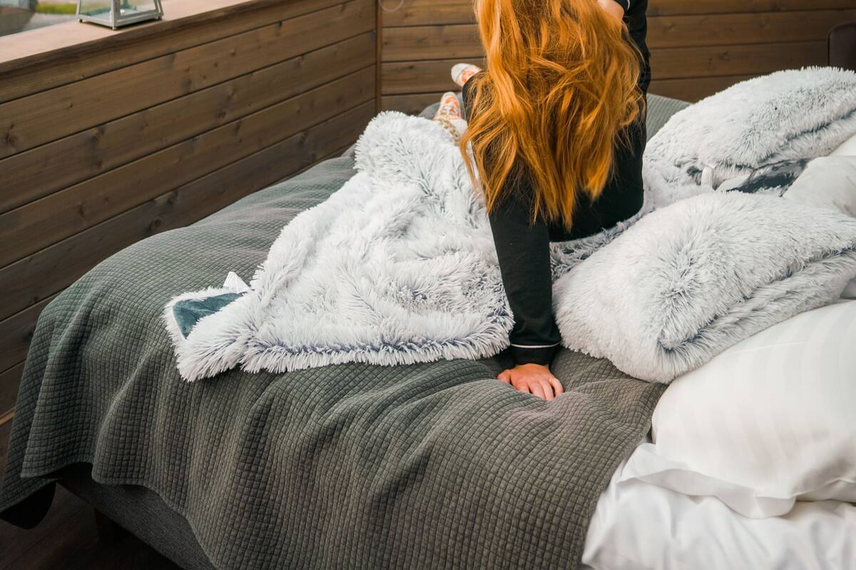 A bedroom with a large window, wooden walls, a bed with a green blanket, and a white fluffy pillow.