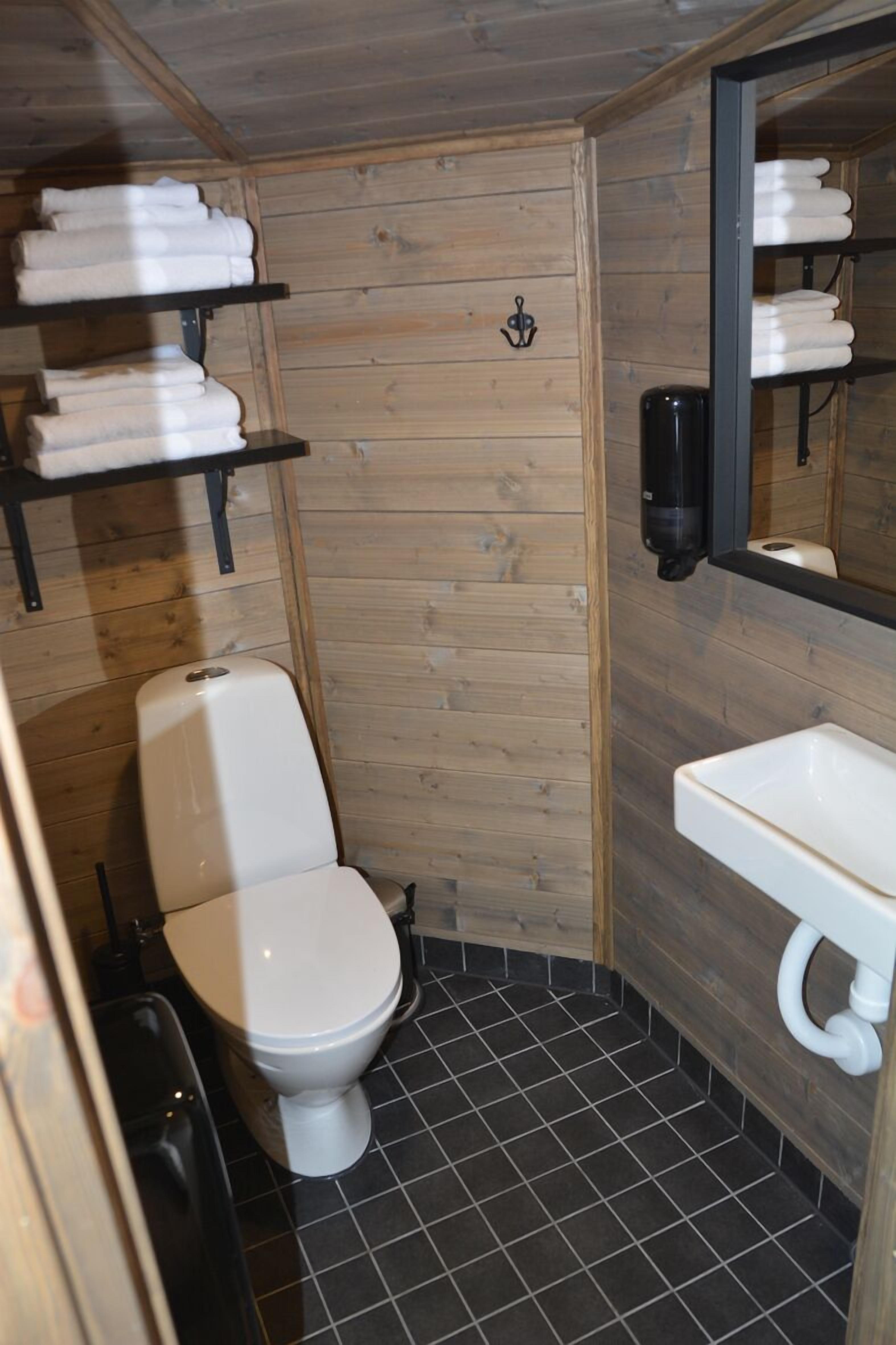 Un baño pequeño de madera con inodoro blanco, lavamanos y repisas con toallas dobladas.