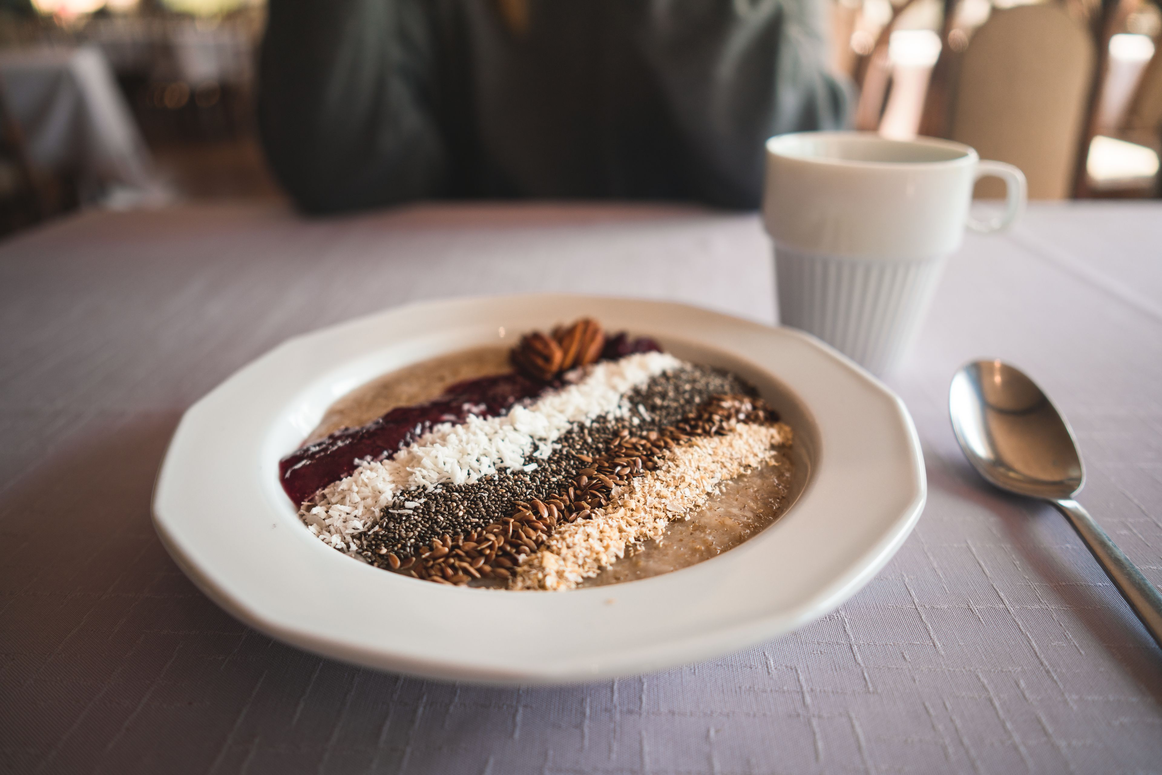 A white plate with a dessert topped with various seeds and a star anise, accompanied by a white mug and a spoon on a table.