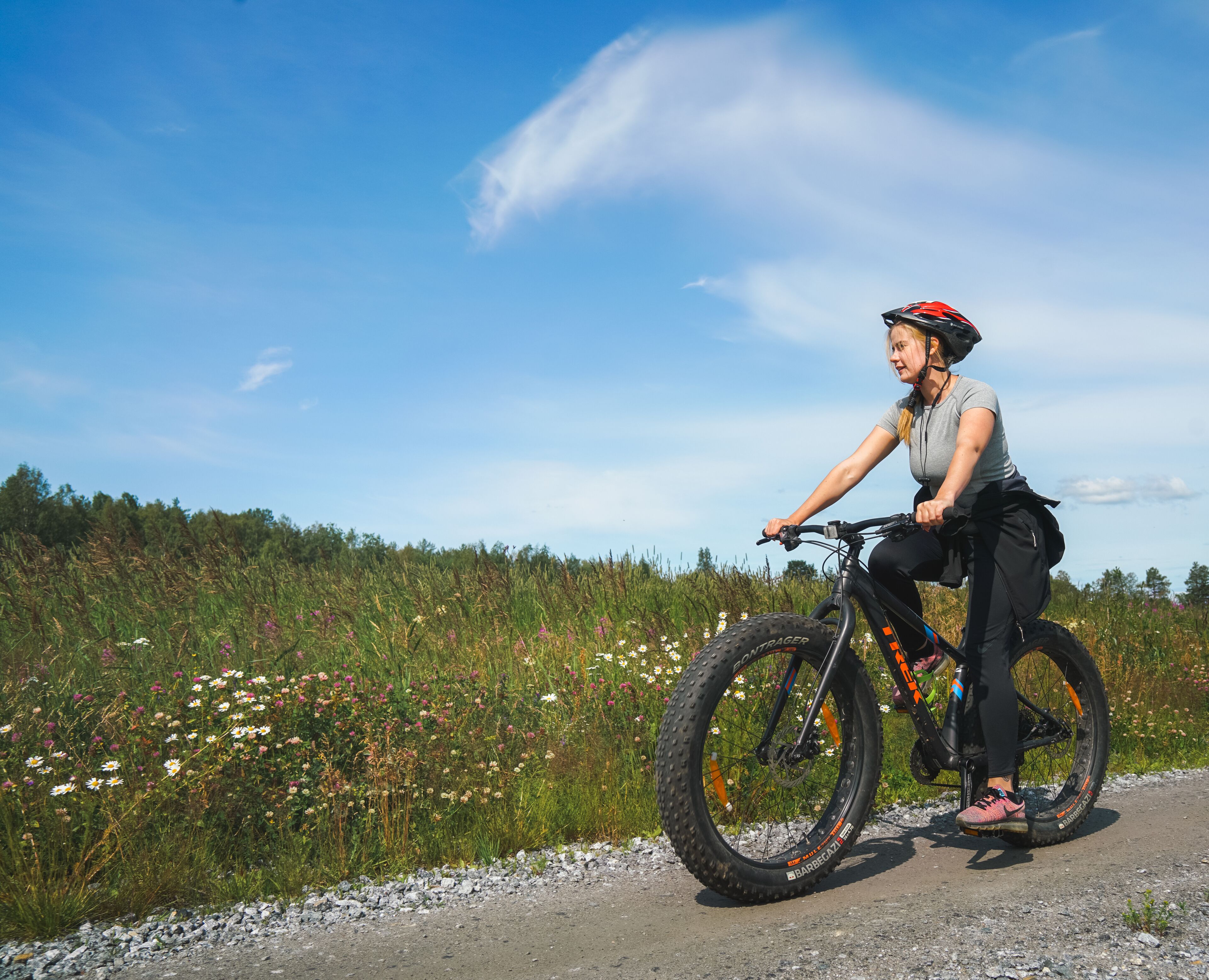 A person riding a mountain bike on a gravel path with wildflowers and trees in the background.