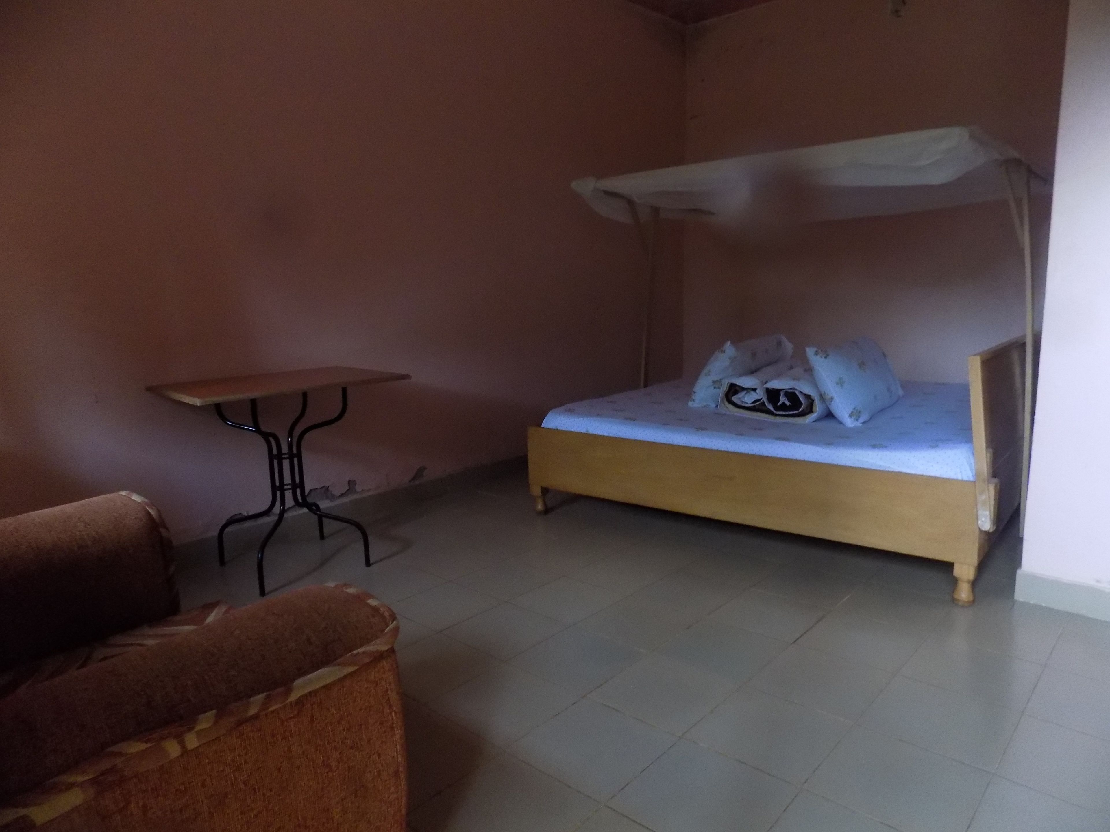 A small, single-bed room with a wooden bed frame, a small table, and a chair.