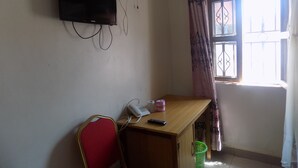 Una habitación con un escritorio de madera, una silla roja, un televisor montado en la pared, una ventana con cortinas y una cesta verde debajo del escritorio.