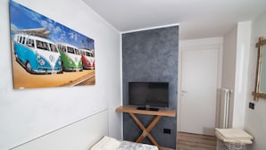 A modern room with a flat-screen TV on a wooden stand, a bed with floral bedding, a small seating area, and a beach-themed painting on the wall. - Da Chris e Lisa 022205-AT-296296 (Trento)