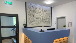 A reception desk with a framed black and white drawing of a town, a potted plant, and a small blue container. - besttime Hotel Monschau (Monschau)
