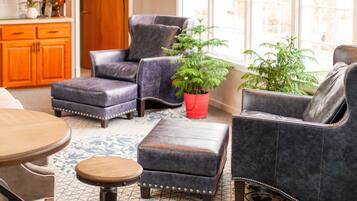 A cozy living room with a leather sofa, ottoman, and a small wooden table. There is a potted plant and a window with a view of the outdoors.