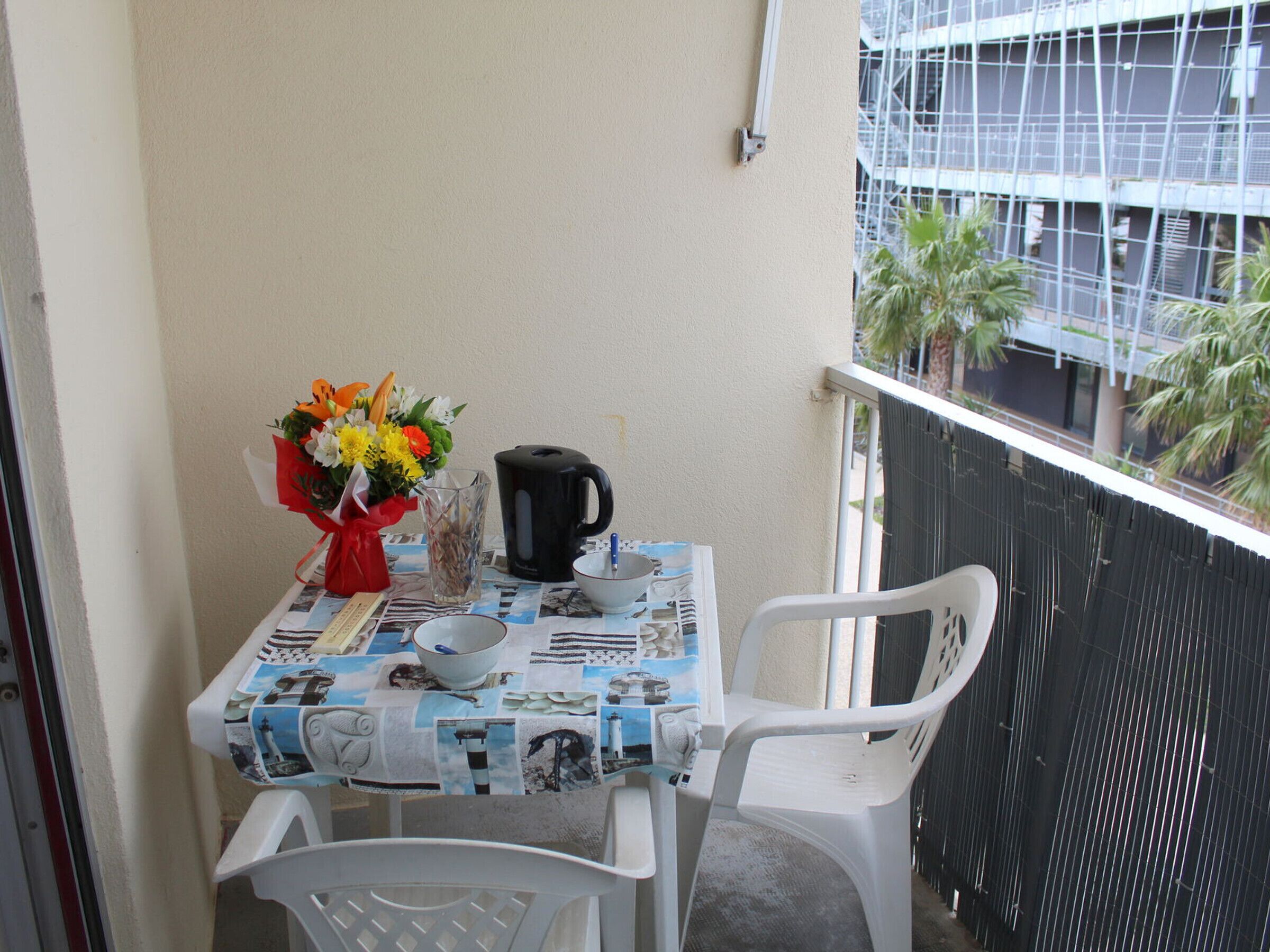 A small balcony with a table set for two, featuring a tablecloth with nautical prints, a vase of flowers, a black coffee pot, and two white plastic chairs.