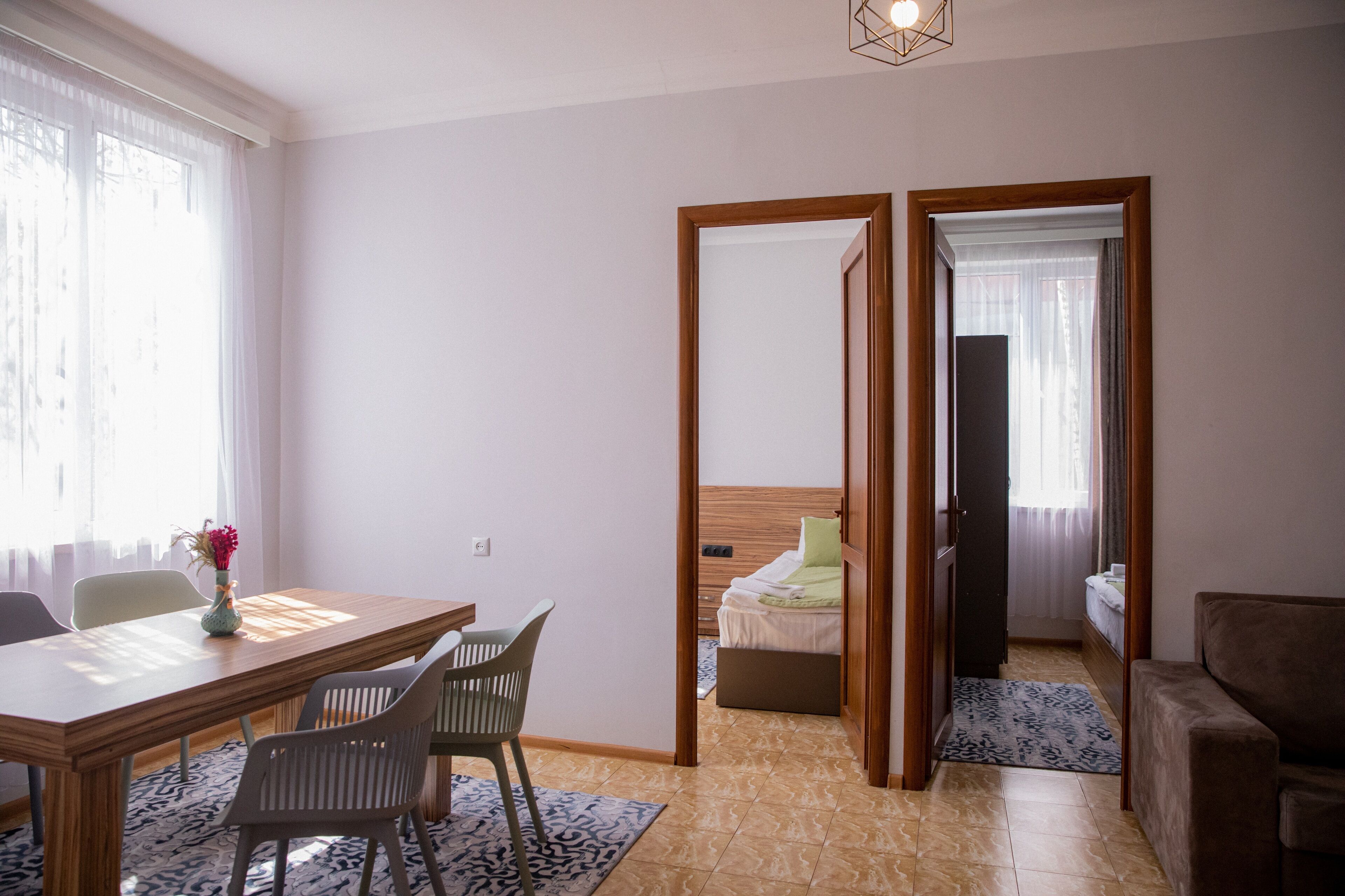 A hotel room with a wooden dining table, two chairs, a bed with a wooden headboard, a sofa, and a small rug.