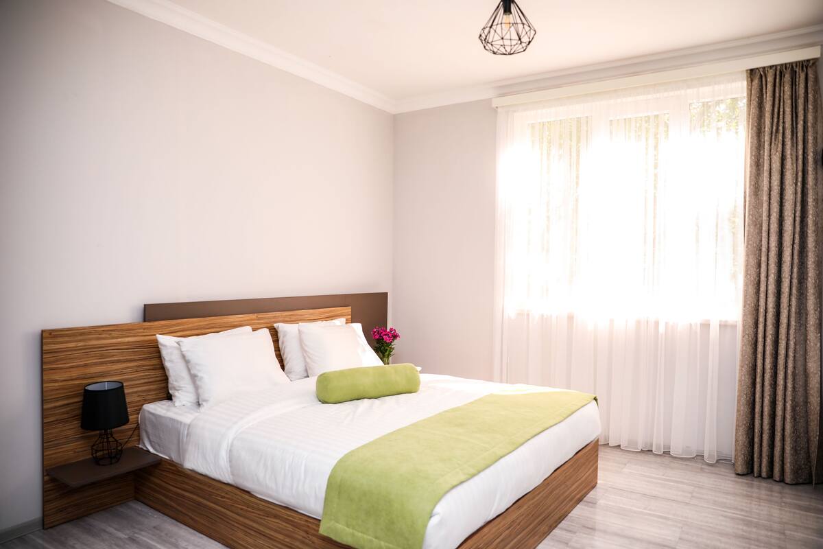 A neatly made bed with white linens and a green throw, a bedside table with a lamp, and a window with sheer curtains and a brown drape.