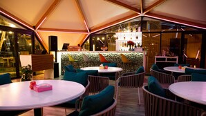 An outdoor dining area with round tables and chairs, a bar with bottles, and a view of the outdoors.