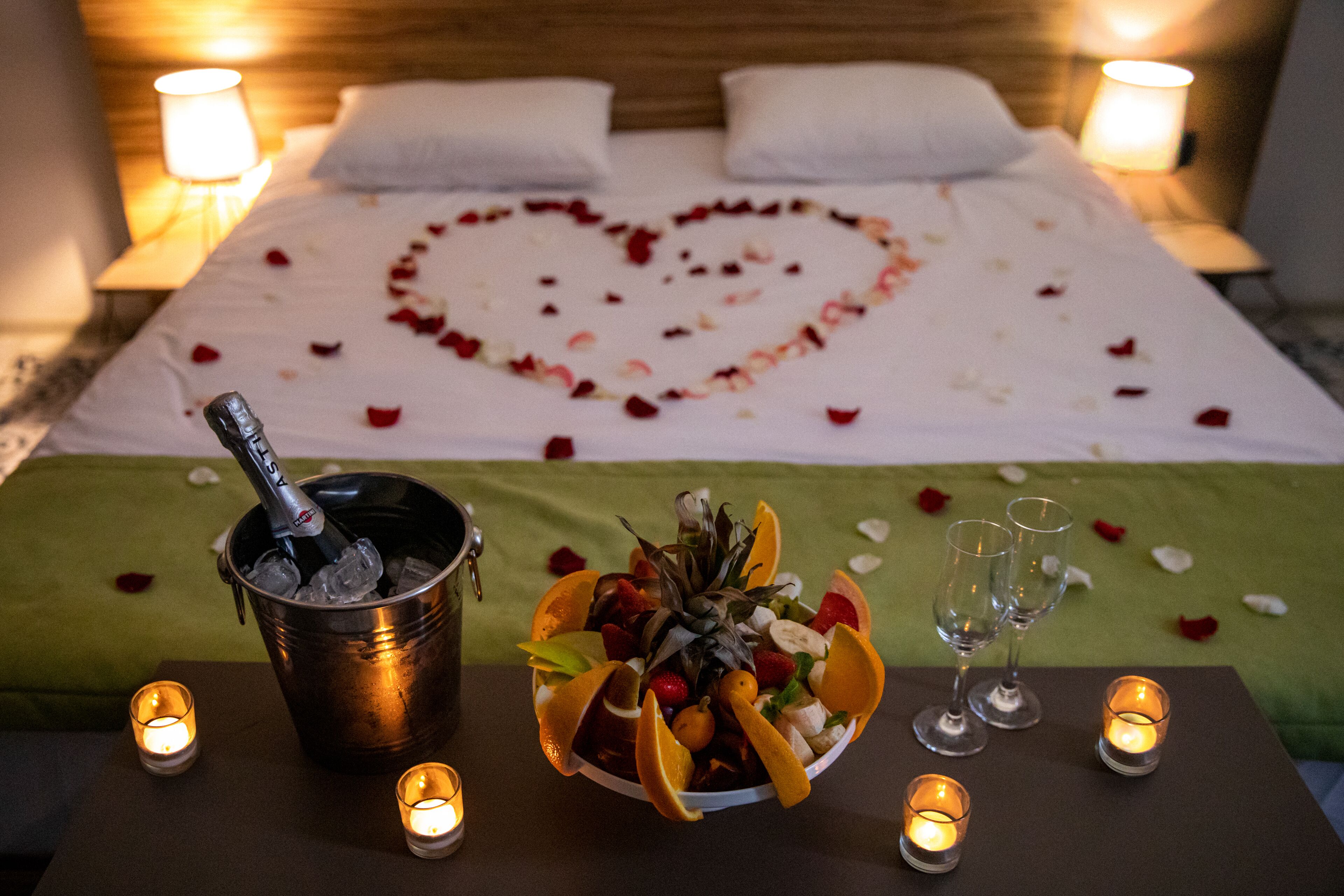 Ein Bett mit einer herzförmigen Anordnung aus Rosenblättern, einem Eiseimer, einer Obstschale und zwei Sektgläsern auf einem Tisch.
