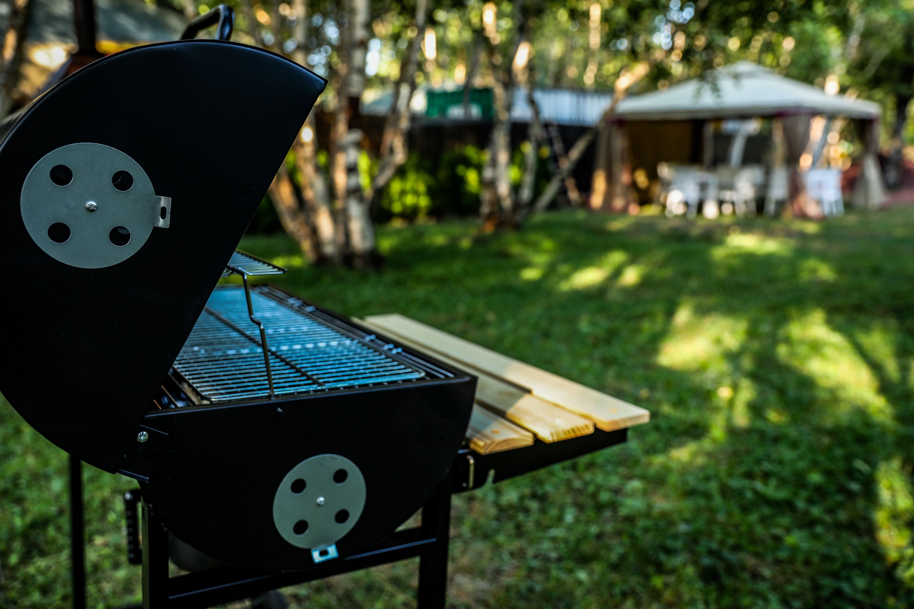 A portable grill with a metal grate, a wooden table, and a grassy area with trees and a tent in the background.