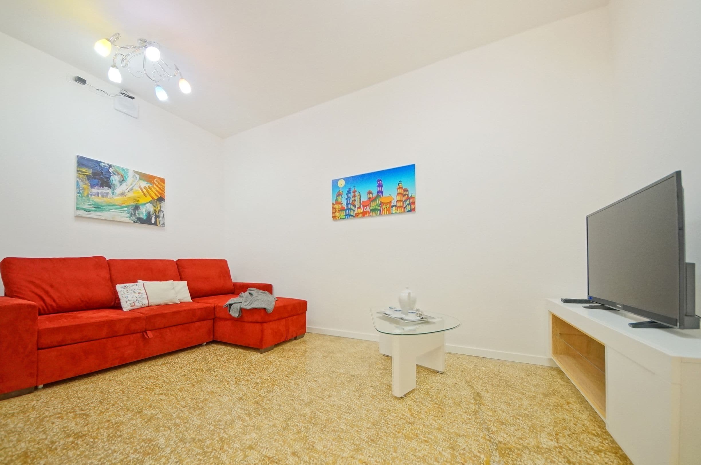 A living room with a red sofa, a glass coffee table, a flat-screen TV on a white stand, and two paintings on the walls.