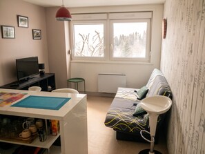 A modern living room with a sofa, a TV, a table with mugs, and a window with a view of trees.