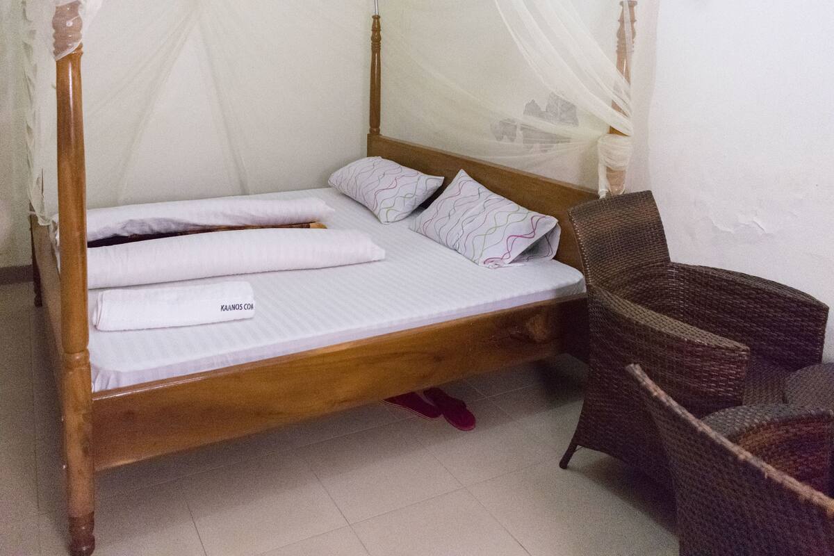 Un simple lit en bois, doté d’un baldaquin, recouvert de draps blancs, et une paire de pantoufles posées sous le pied de lit.