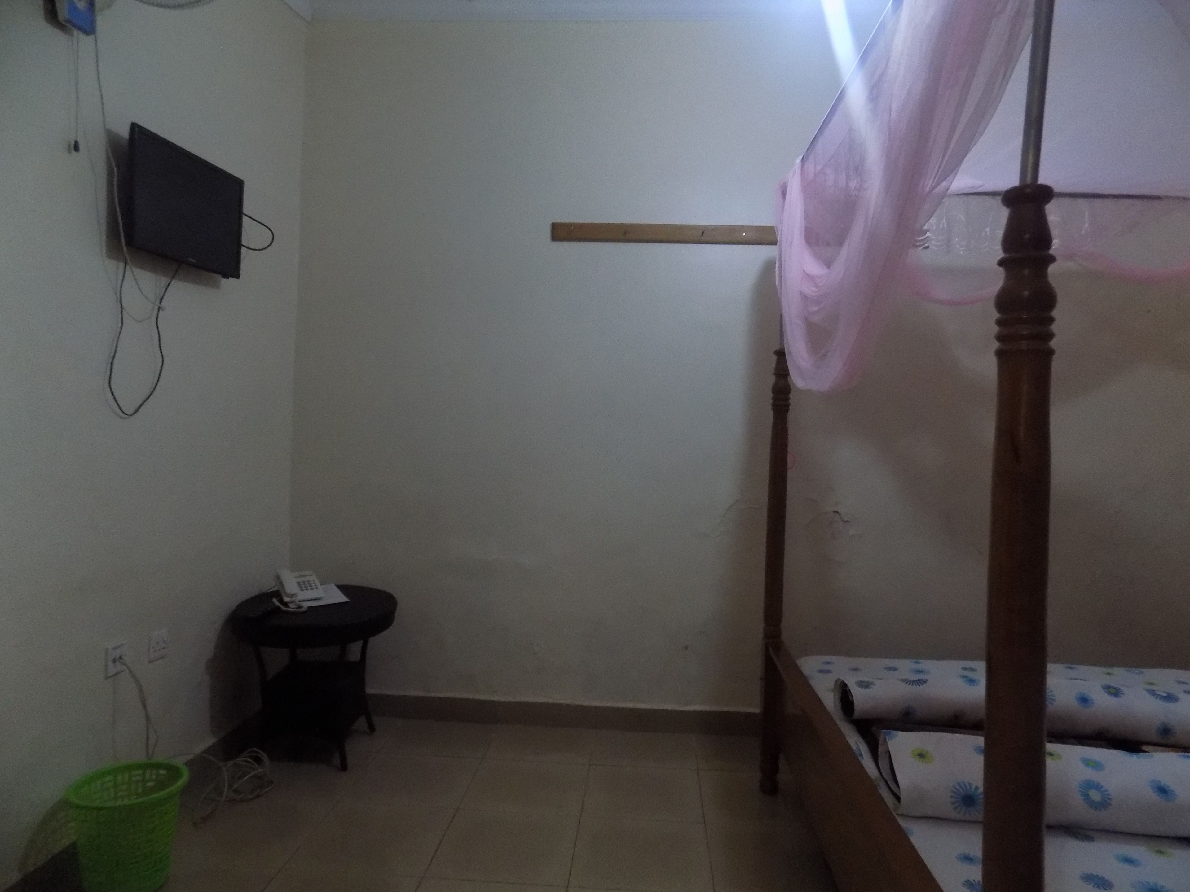 A small, dimly lit room with a single bed, a small table, and a television mounted on the wall.