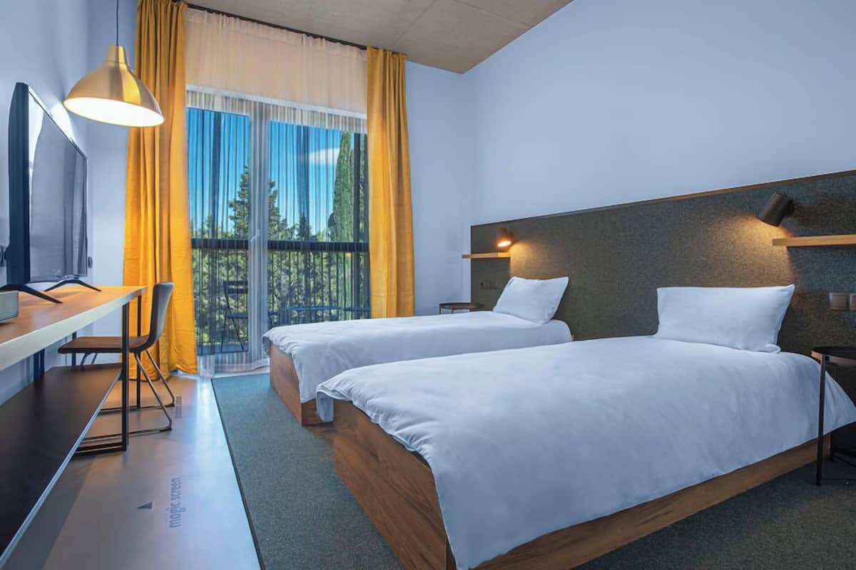 A modern hotel room with two beds, a desk, and a balcony with yellow curtains.