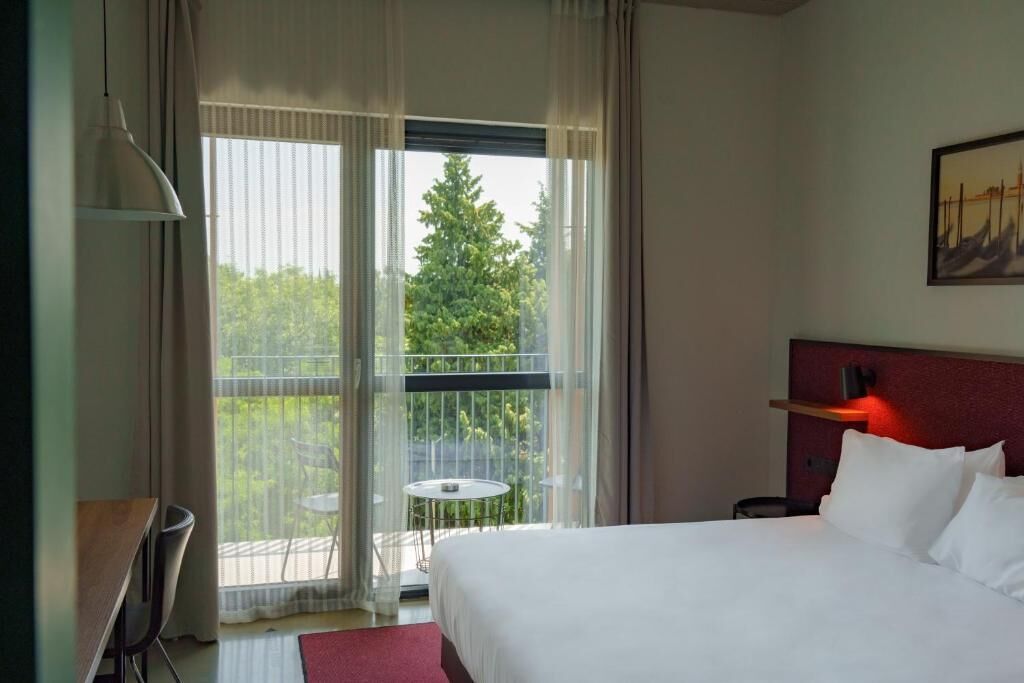 A hotel room with a bed, a desk, a chair, a small table, and a balcony with a view of trees.