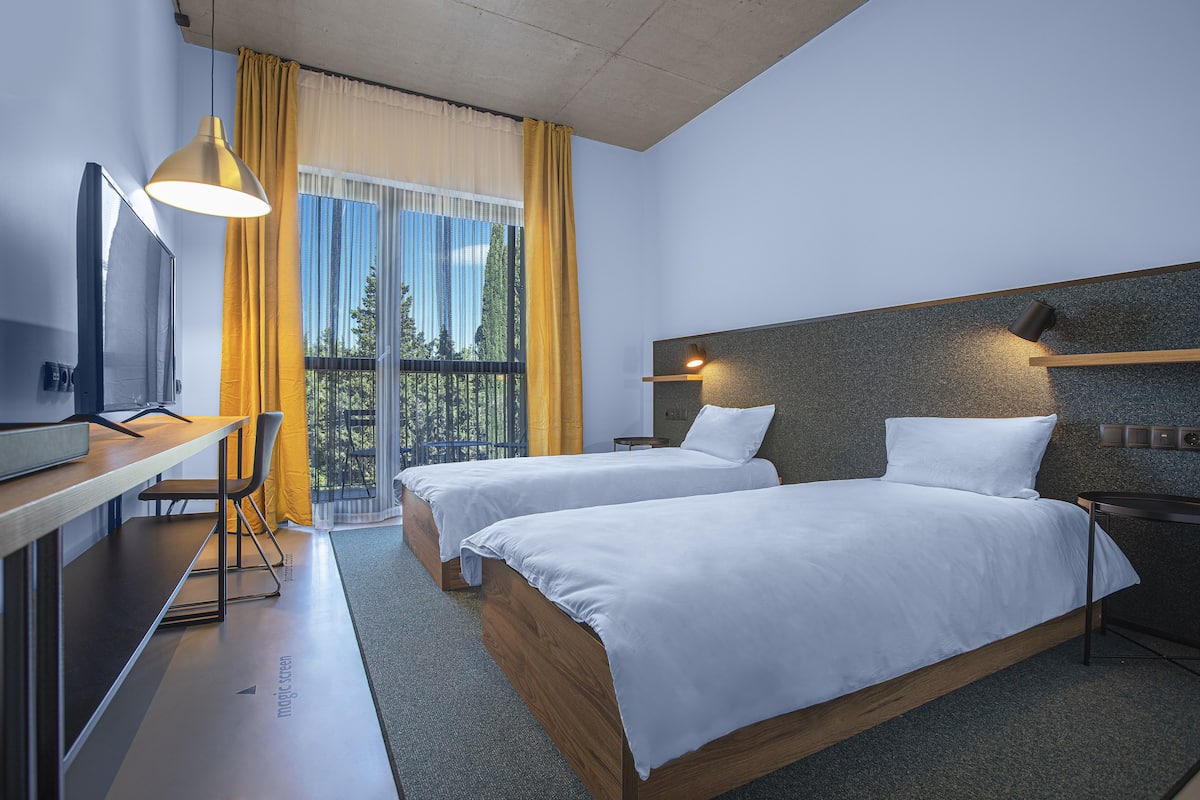 A modern hotel room with two beds, a desk, and a balcony with yellow curtains.