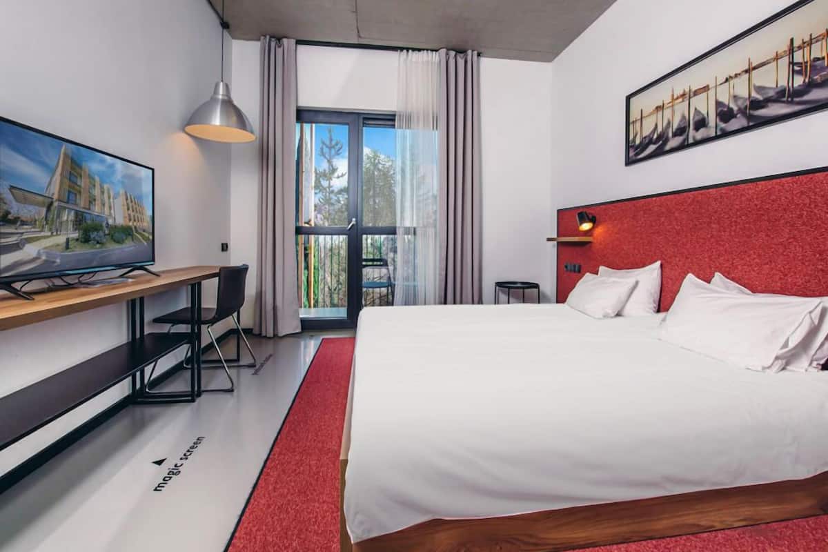 A modern hotel room with a large bed, a desk, a television, and a balcony with curtains.