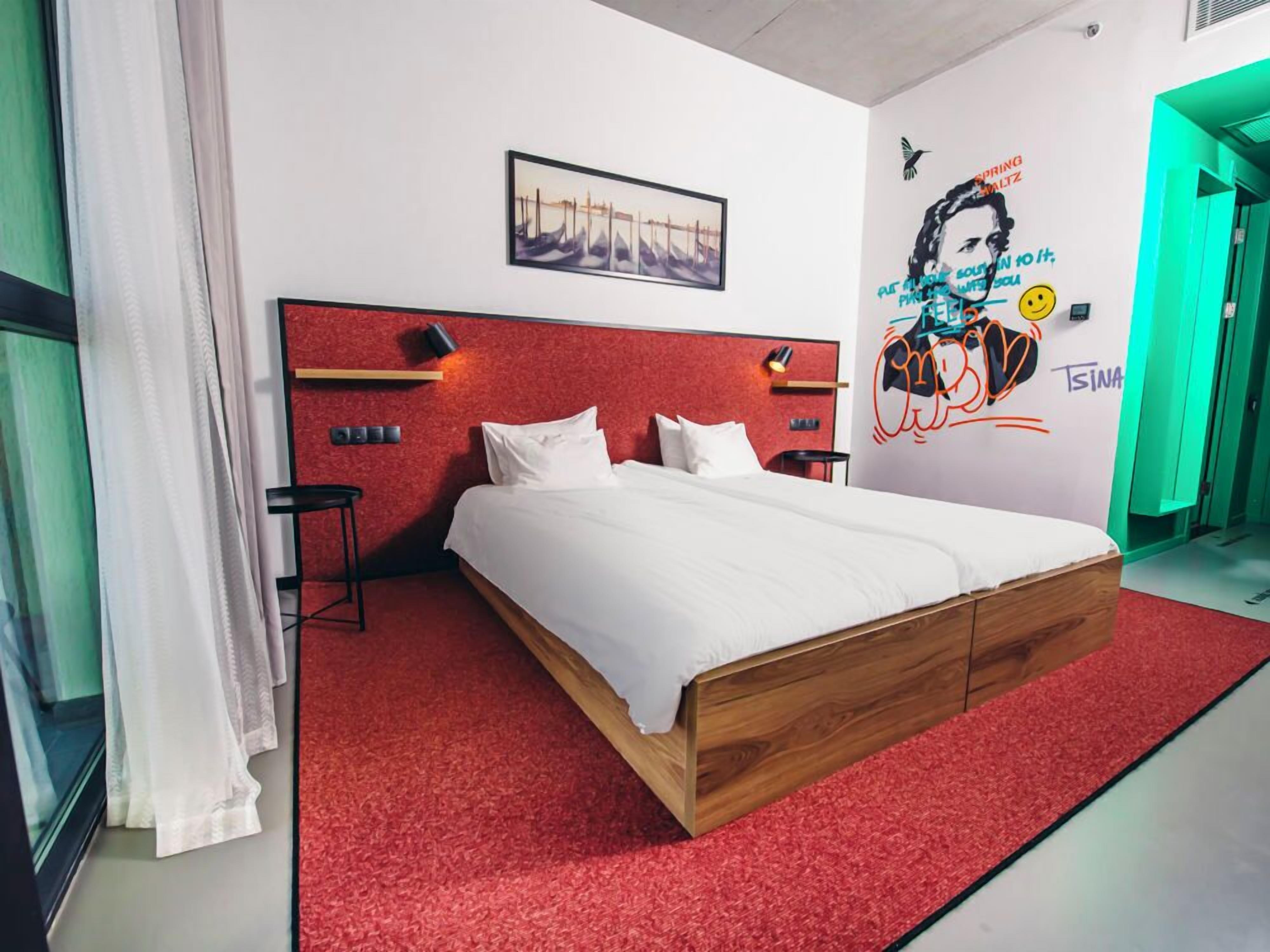 Une chambre d’hôtel moderne avec un tapis rouge, un cadre de lit en bois, du linge de lit blanc, un tableau encadré au mur et une fresque murale avec du texte et un visage souriant.