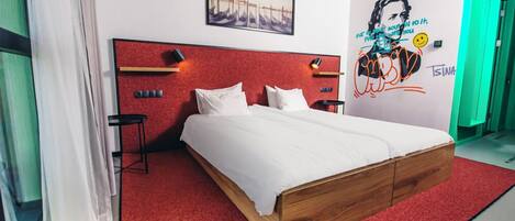 Une chambre d’hôtel moderne avec un tapis rouge, un cadre de lit en bois, du linge de lit blanc, un tableau encadré au mur et une fresque murale avec du texte et un visage souriant.