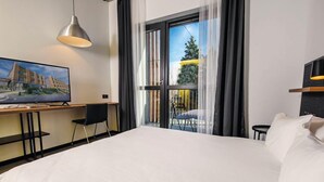 A hotel room with a large bed, a desk with a chair, a TV, and a balcony with a view of a building and trees. - Park Hotel Tsinandali Estate (Telavi)