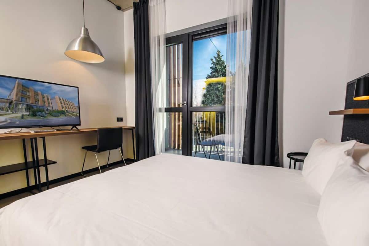 A hotel room with a large bed, a desk with a chair, a TV, and a balcony with a view of a building and trees.