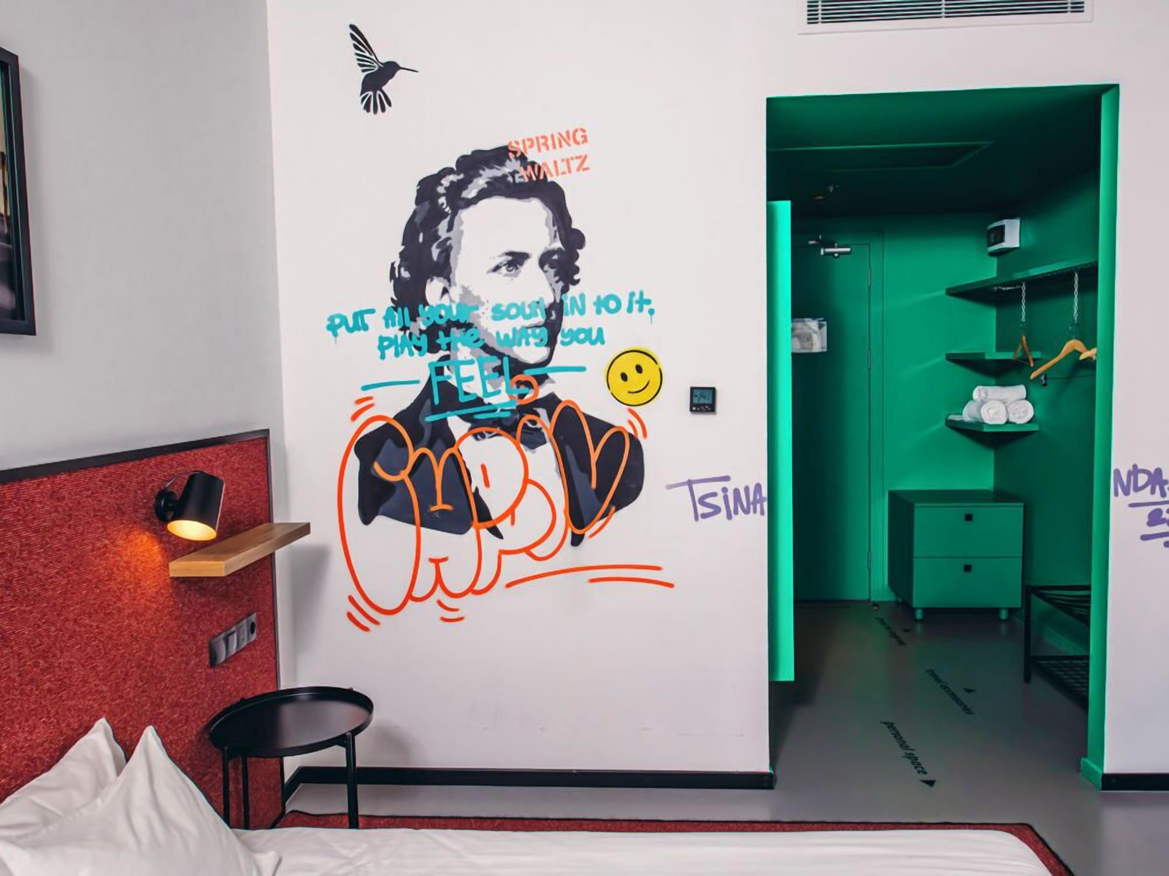 Una habitación de hotel con un mural, una cama, una mesita de noche y una pared verde.